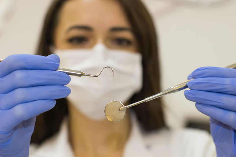 A woman in a white coat, blue latex gloves, and a medical mask is holding up a dental scraper and a dental mouth mirror.